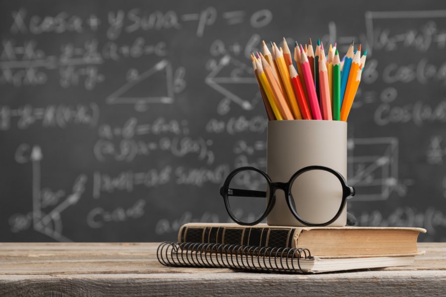 A book, pencils, and glasses on a table in front of a chalkboard.